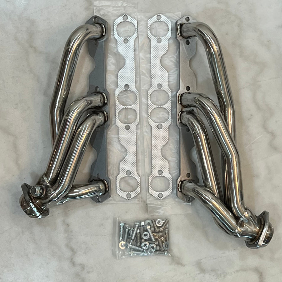 Polished stainless steel exhaust headers for vehicles, displayed on a marble surface, including gaskets and mounting hardware.